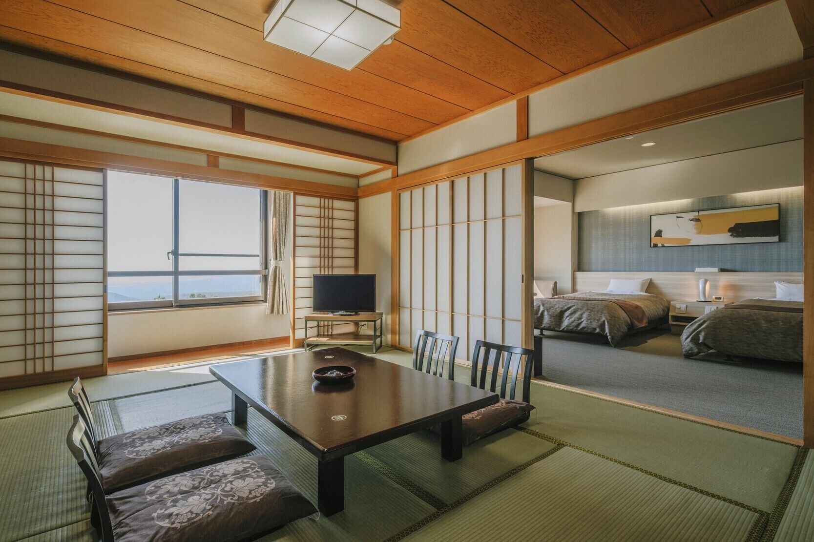 A traditional Japanese-style room with tatami flooring, a low table with cushions, a television, and a bed with futons.