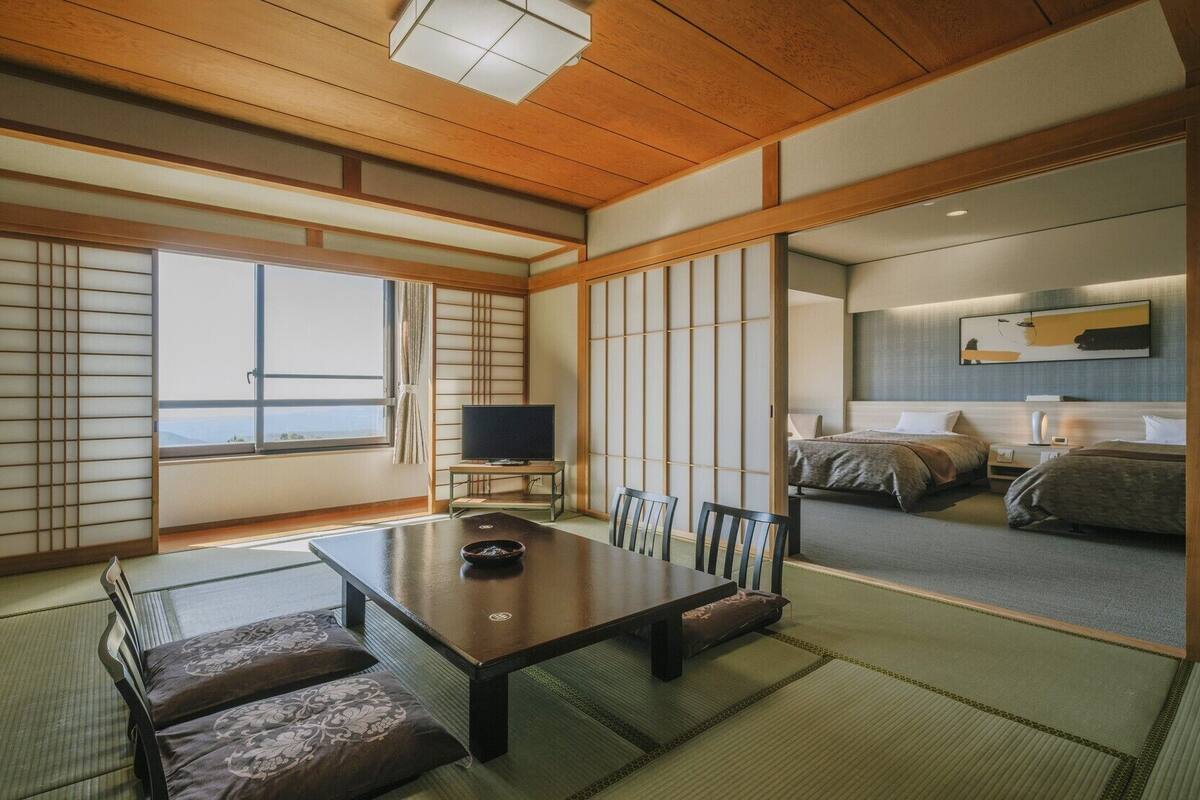 Una habitación tradicional japonesa con suelo de tatami, una mesa baja con cojines, televisión y una cama con futones.