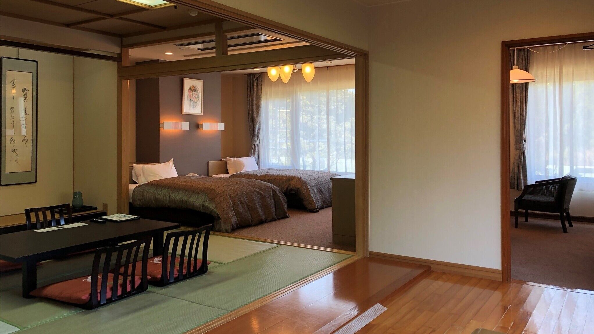 A hotel room with a traditional Japanese interior, featuring tatami flooring, a low table with cushions, a bed with a patterned bedspread, and a sliding door leading to another room.