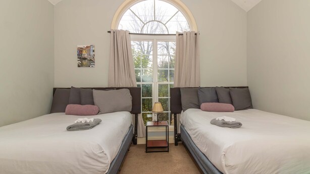A room with two beds, a window with curtains, a small table with a lamp, and a painting on the wall. - Creekside Stay Near Fort Benning Sleeps 12! (Columbus)
