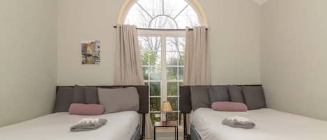 A room with two beds, a window with curtains, a small table with a lamp, and a painting on the wall.