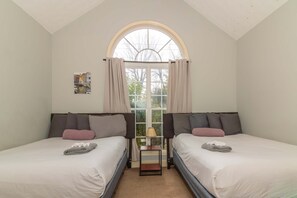 Quarto com duas camas, janela com cortinas, uma mesinha com abajur e um quadro na parede.