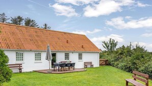 Une maison blanche avec un toit rouge, une terrasse avec une table et des chaises, et une cour couverte d’herbe.