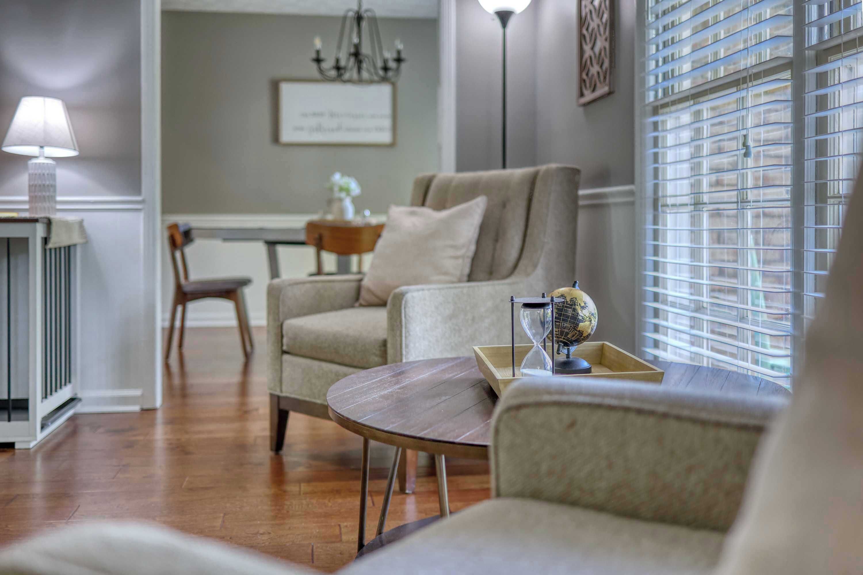 Un salon avec un canapé, une petite table et une lampe. Il y a une fenêtre avec des stores et un lustre au mur.