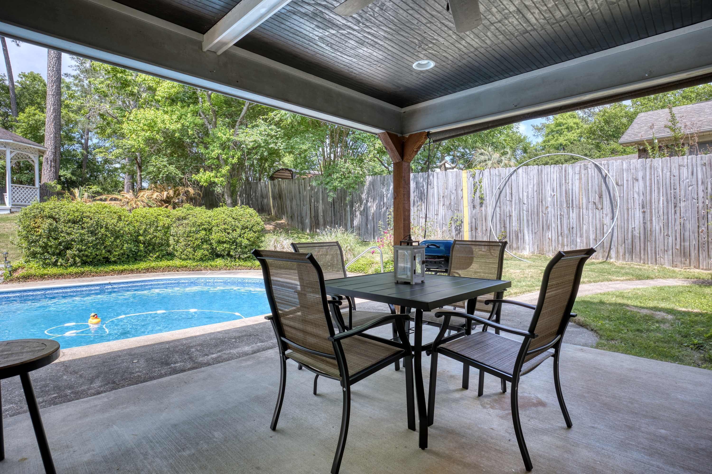 A covered patio area with a pool, outdoor furniture, and a wooden fence in the background.