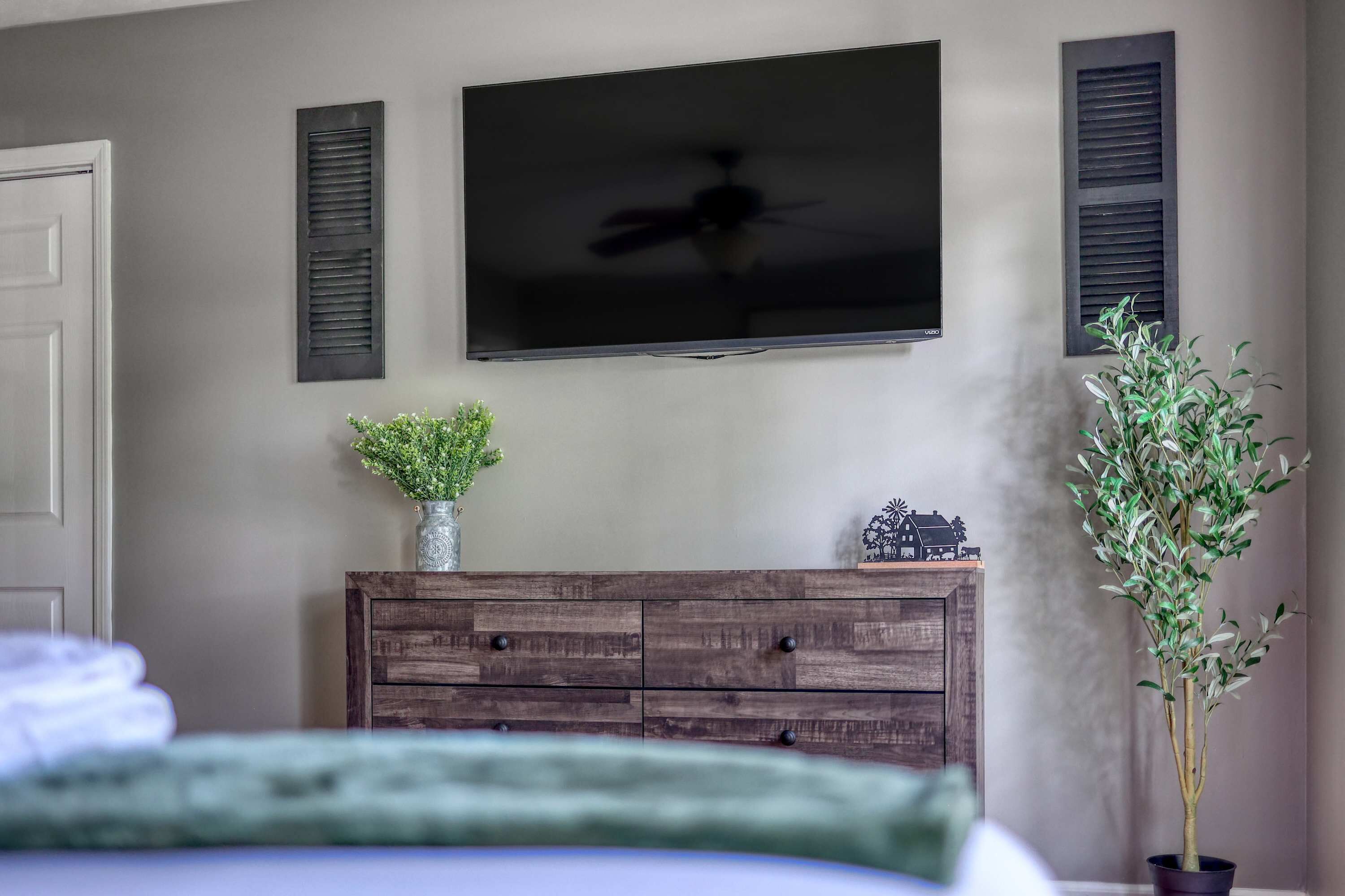 A bedroom with a bed, a dresser, a television mounted on the wall, and two potted plants.