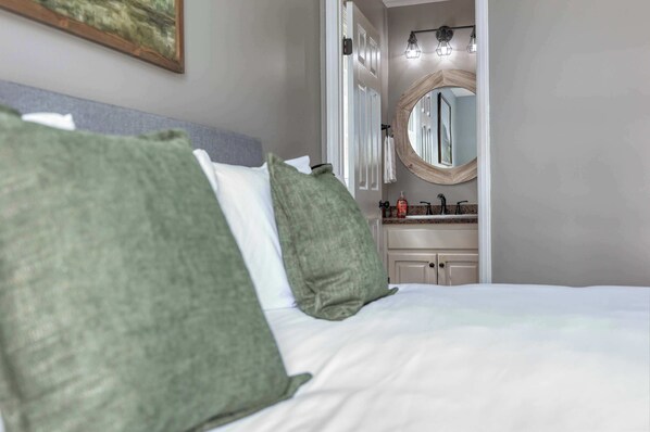 A hotel room with a bed, green pillows, a bathroom with a sink and mirror, and a painting on the wall.