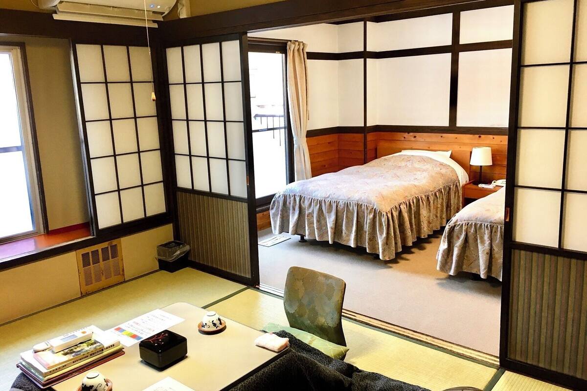 Ein traditioneller japanischer Raum mit Tatami-Bodenbelag, einer Futon-Liege, einem Schreibtisch mit Büchern und einem Teeset sowie einer raumteilenden Shoji-Schiebetür.