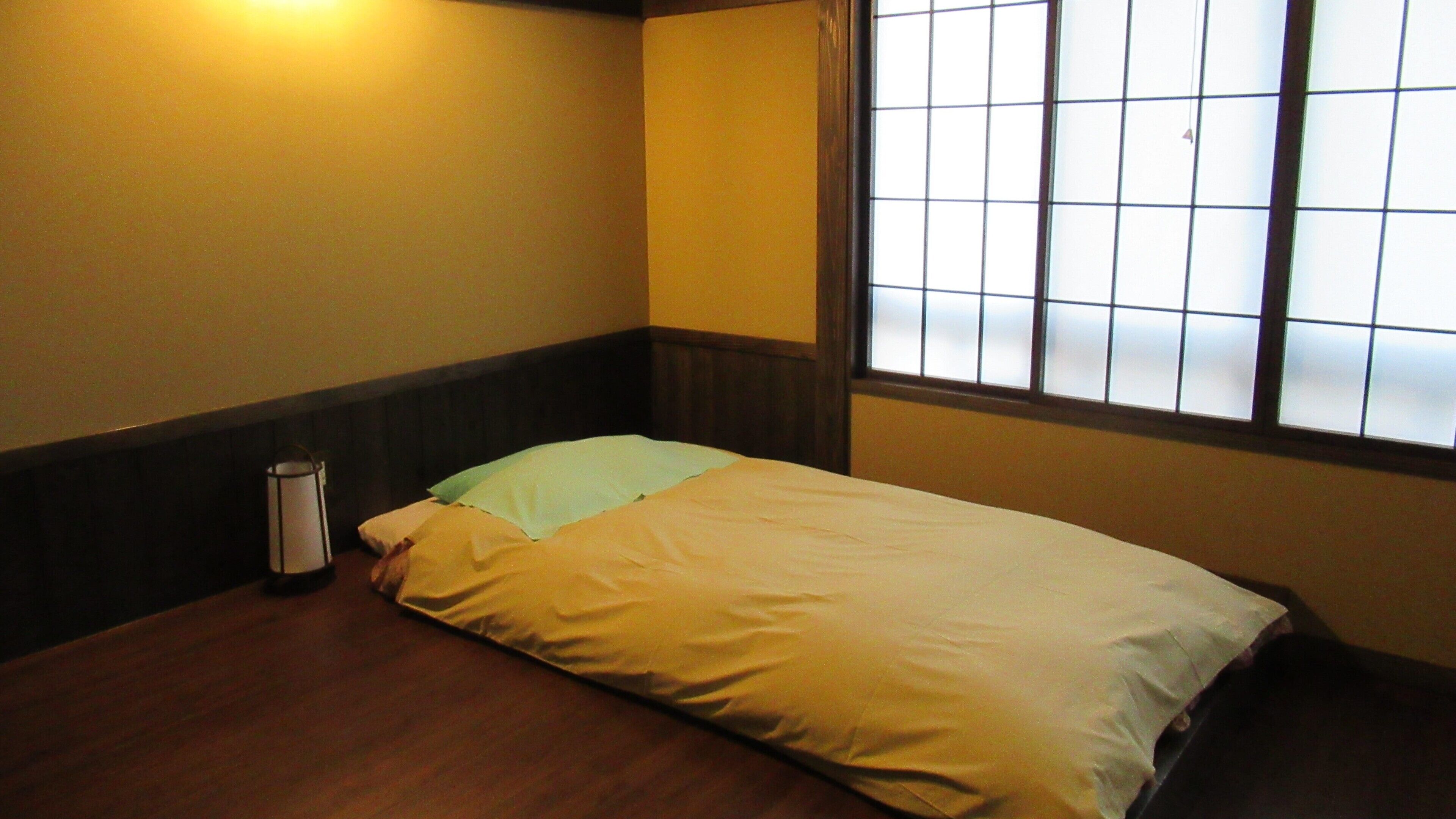 Una habitación con una cama, una mesita de noche, una ventana con persianas y una lámpara de pared.