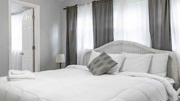 A neatly made bed with a white duvet and patterned pillows, a bedside lamp, and a door to the left. - Historic Edgewood Home: Modern Charm, (Columbus)