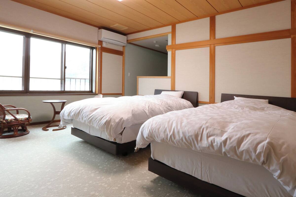A hotel room with two beds, a small table, a chair, and a large window.