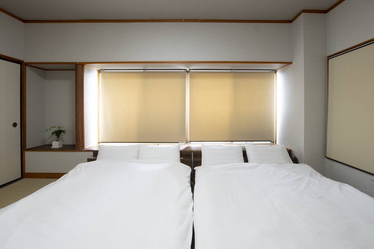 A hotel room with two beds, a window with blinds, a small bedside table with a plant, and a wooden ceiling.