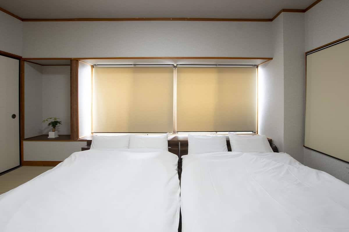 A hotel room with two beds, a window with blinds, a small bedside table with a plant, and a wooden ceiling.