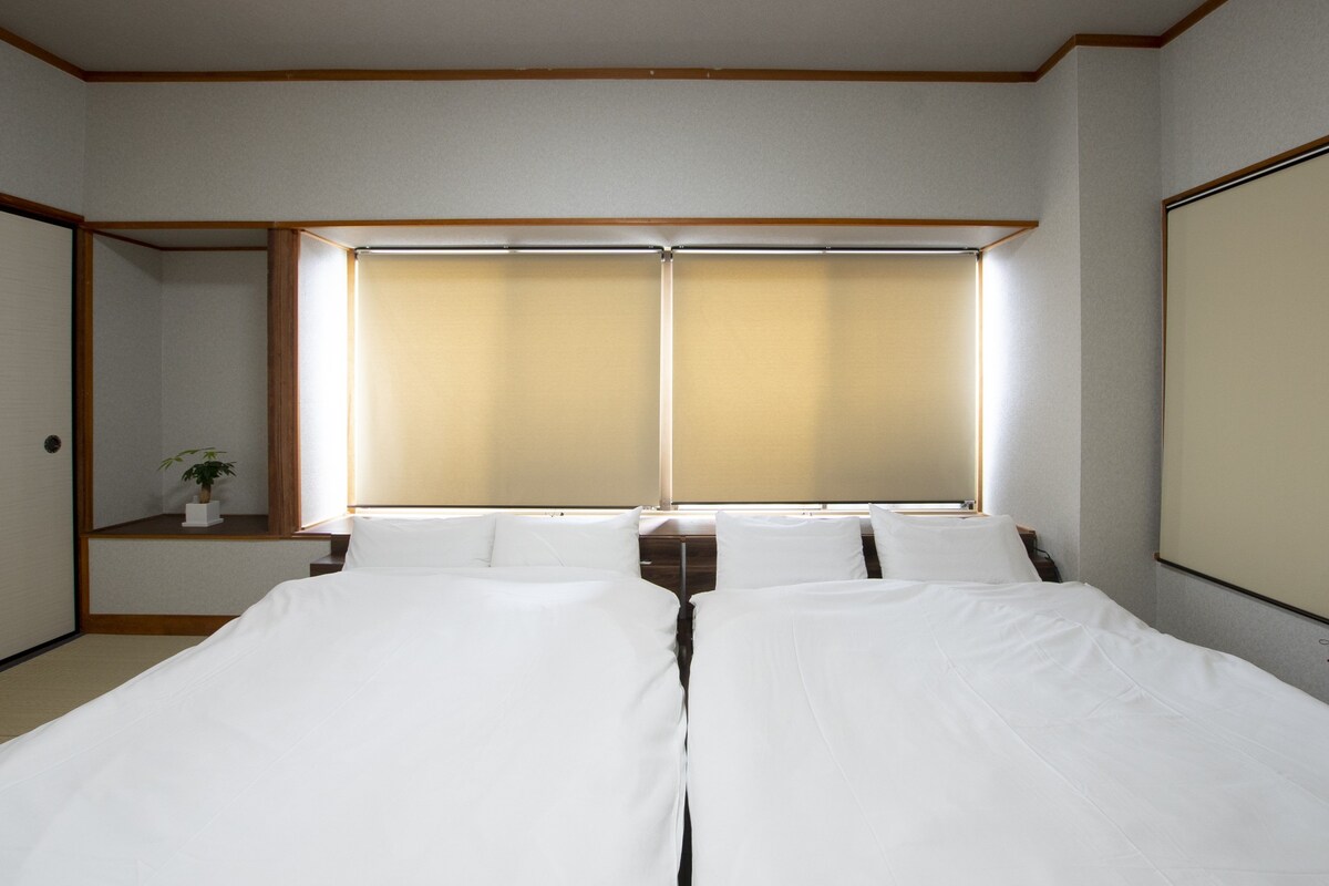 A hotel room with two beds, a window with blinds, a small bedside table with a plant, and a wooden ceiling.