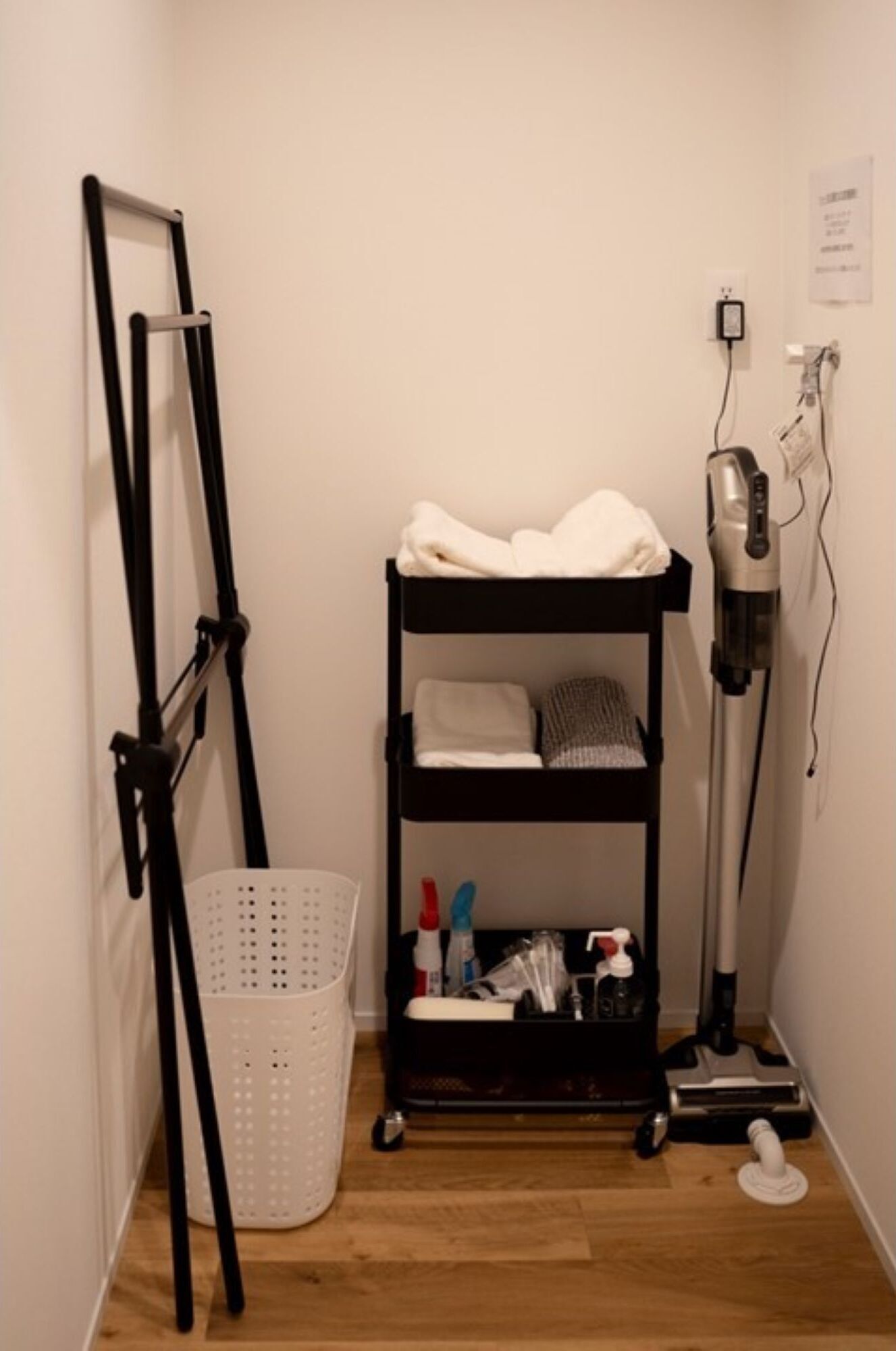 A corner room with a black shelf unit containing a hairdryer, a vacuum cleaner, and toiletries. A white laundry basket is on the floor.