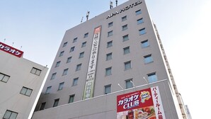 Un hôtel à plusieurs étages, doté d’une enseigne bien visible indiquant « APA HOTEL » et d’une grande banderole verticale faisant la publicité du « DAD Club » et du « DAM Resort ».