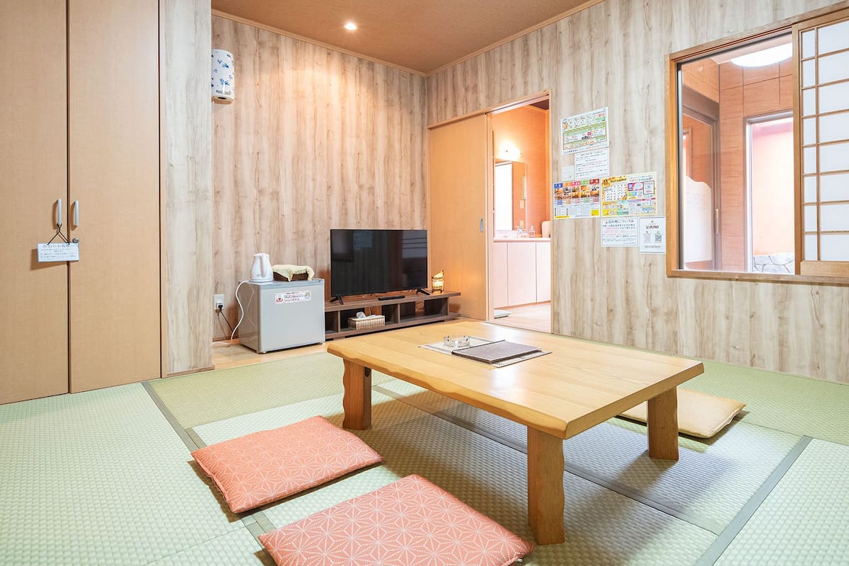 Ein traditioneller japanischer Raum mit Tatami-Boden, einem niedrigen Holztisch, einem Fernseher und einer kleinen Küchenzeile.