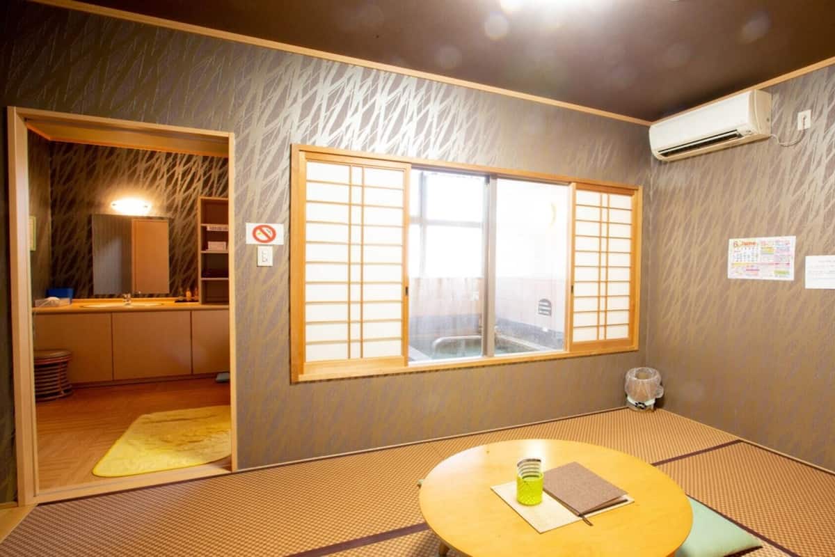 Ein traditioneller japanischer Raum mit Tatami-Bodenbelag, einem Shoji-Fenster, einem kleinen Tisch mit einem grünen Behälter und einem Notizbuch sowie einer wandmontierten Klimaanlage.
