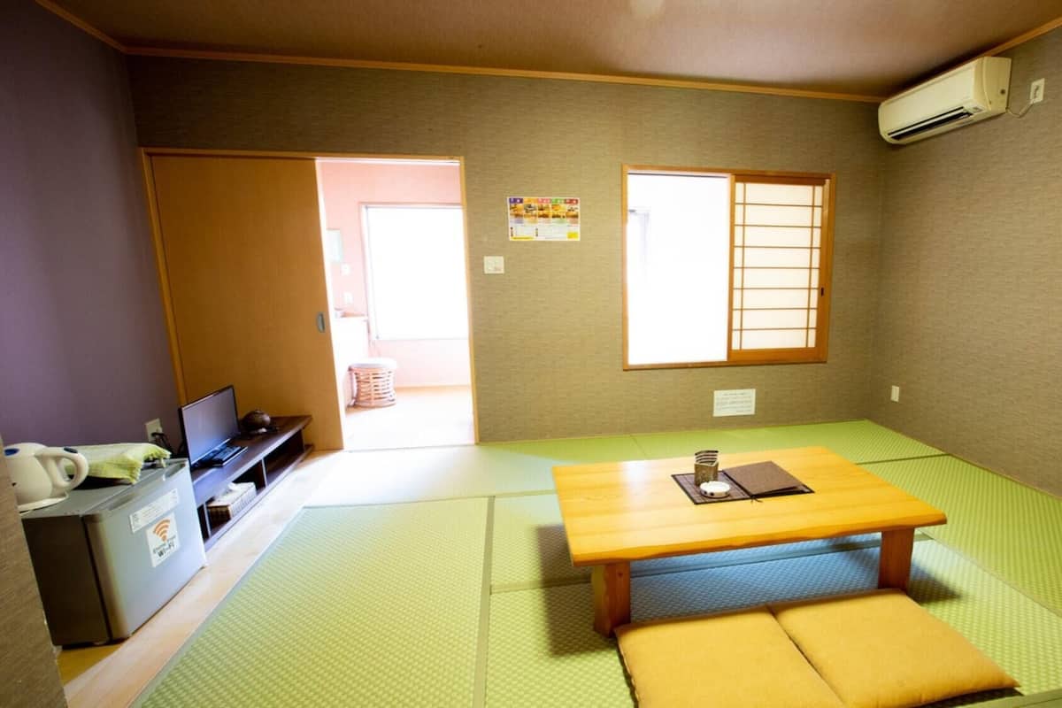 Ein Zimmer mit Tatami-Boden, einem Holztisch, einer Couch, einem Schreibtisch mit Computer und einer Klimaanlage.