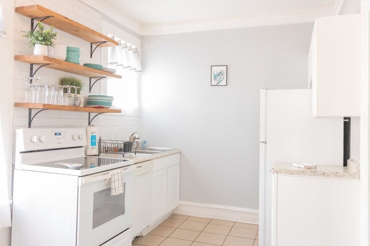 A kitchen with white cabinetry, a white stove, and a sink. There are wooden shelves above the stove holding dishes and a plant.