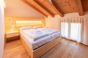 Ein hölzernes Bett in einem Raum mit schrägen Wänden und einem Fenster mit durchsichtigen Vorhängen.