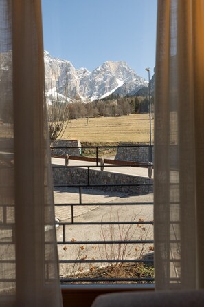 A view through a window with a stone wall and a metal fence, leading to a paved area with benches and a mountain range in the background. - 32 Casa Ladinia 2, comfortable and modern (San Vito di Cadore)