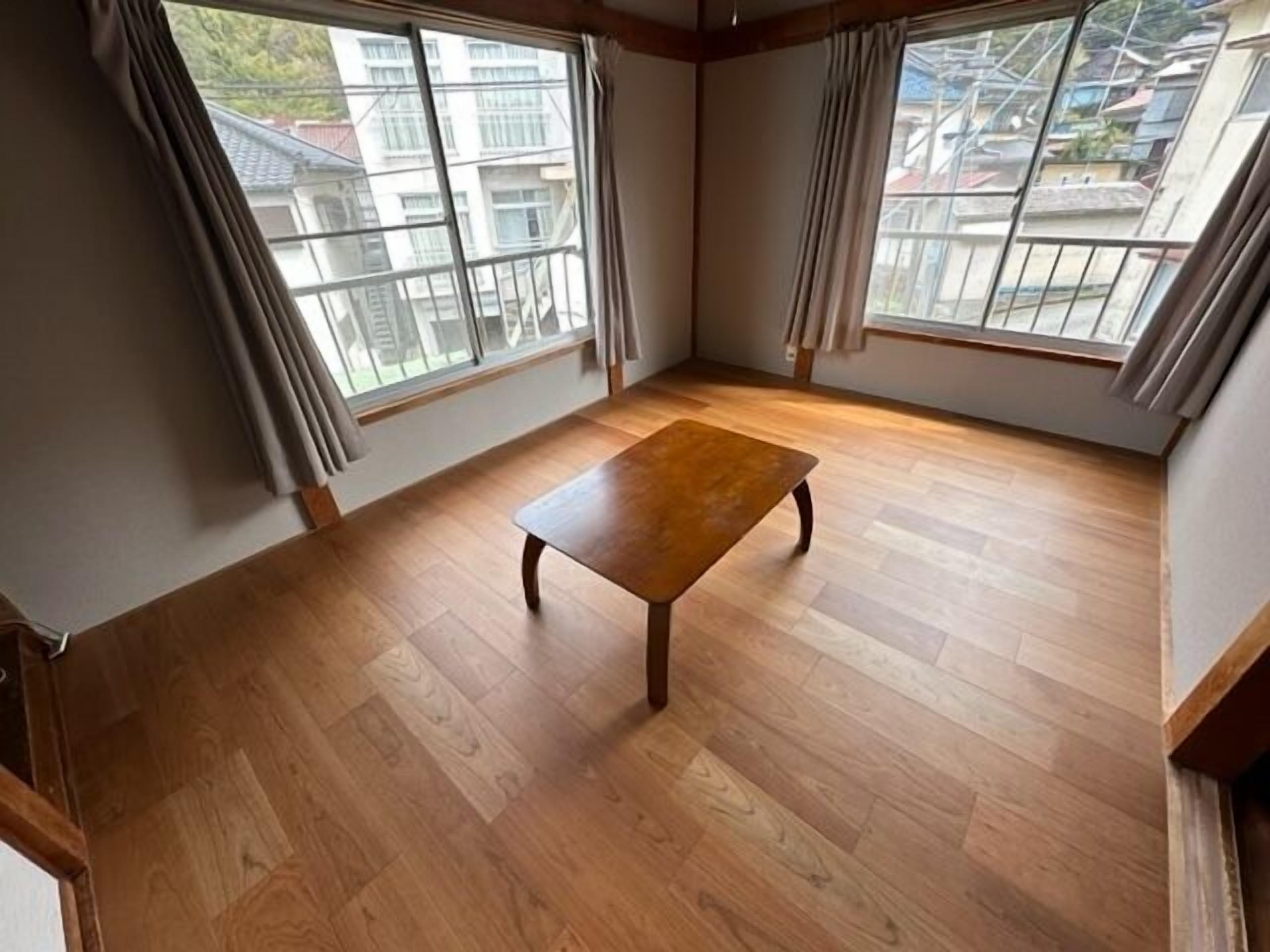 Ein Zimmer mit Holzboden, einem zentralen Tisch und großen Fenstern mit Vorhängen.