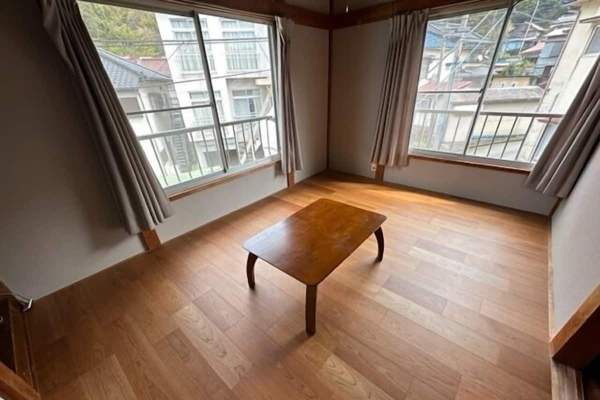 Una habitación con suelo de madera, una mesa central y amplios ventanales con cortinas.