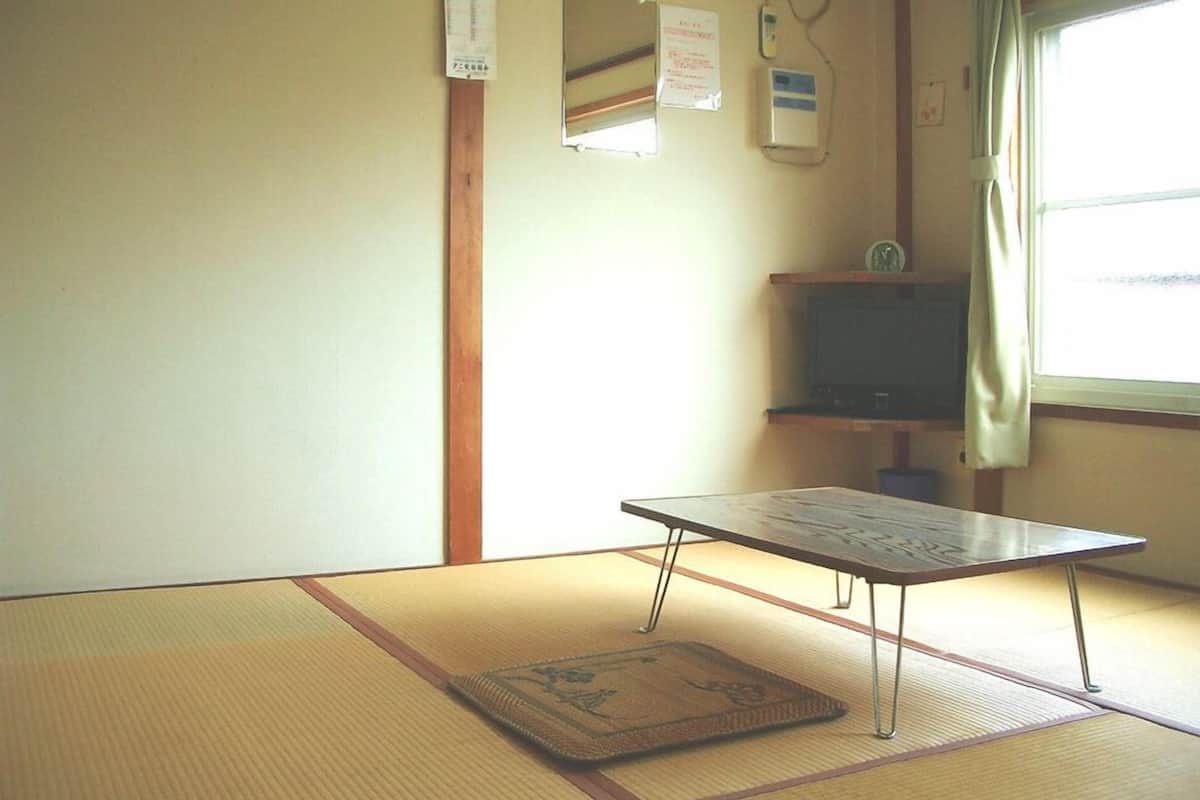 Una habitación con esteras de tatami, una mesa baja, un televisor sobre un mueble y una ventana con cortinas.