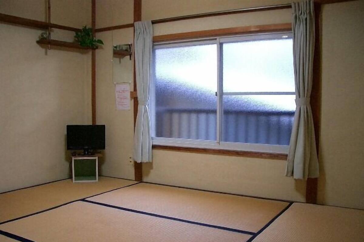 Una habitación con suelo de tatami, un televisor, una ventana con cortinas y una pequeña estantería con una planta.