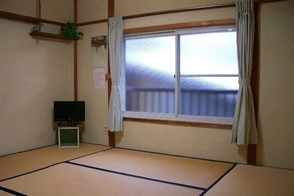 A tatami mat room with a television, a window with curtains, and a small shelf with a plant.