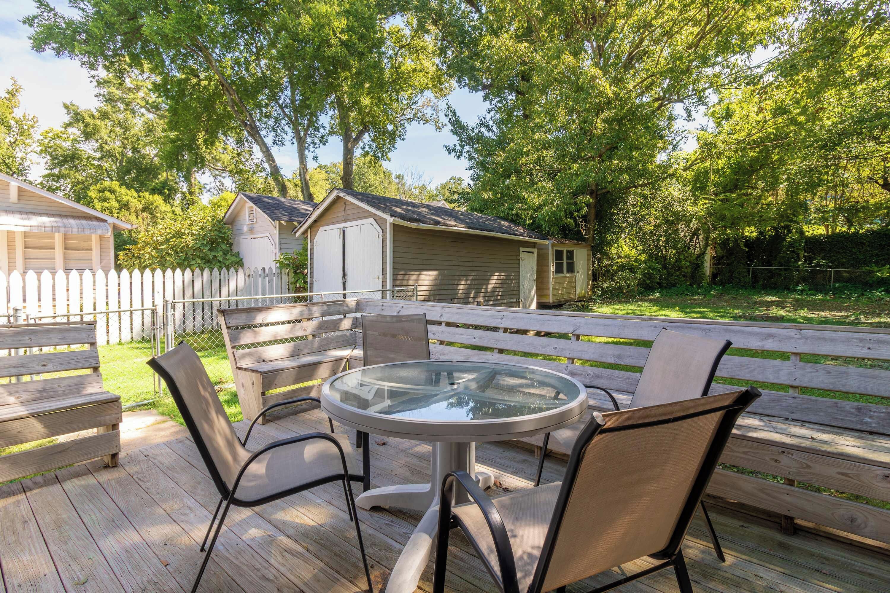 Een terras buiten met een ronde glazen tafel en vier stoelen, omgeven door een houten terras en een witte picket fence.