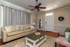 A living room with a beige sofa, a coffee table, a patterned rug, a ceiling fan, and a door with a wreath. - Historic Parkside Home w/ Fenced Yard, (Columbus)