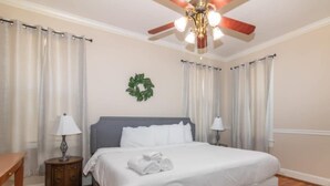 Un lit bien fait, avec du linge de lit blanc, un ventilateur de plafond et une petite table avec une lampe.
