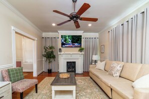 Un salon avec un ventilateur de plafond, un téléviseur fixé au-dessus d’une cheminée, un canapé, une table basse et un fauteuil.