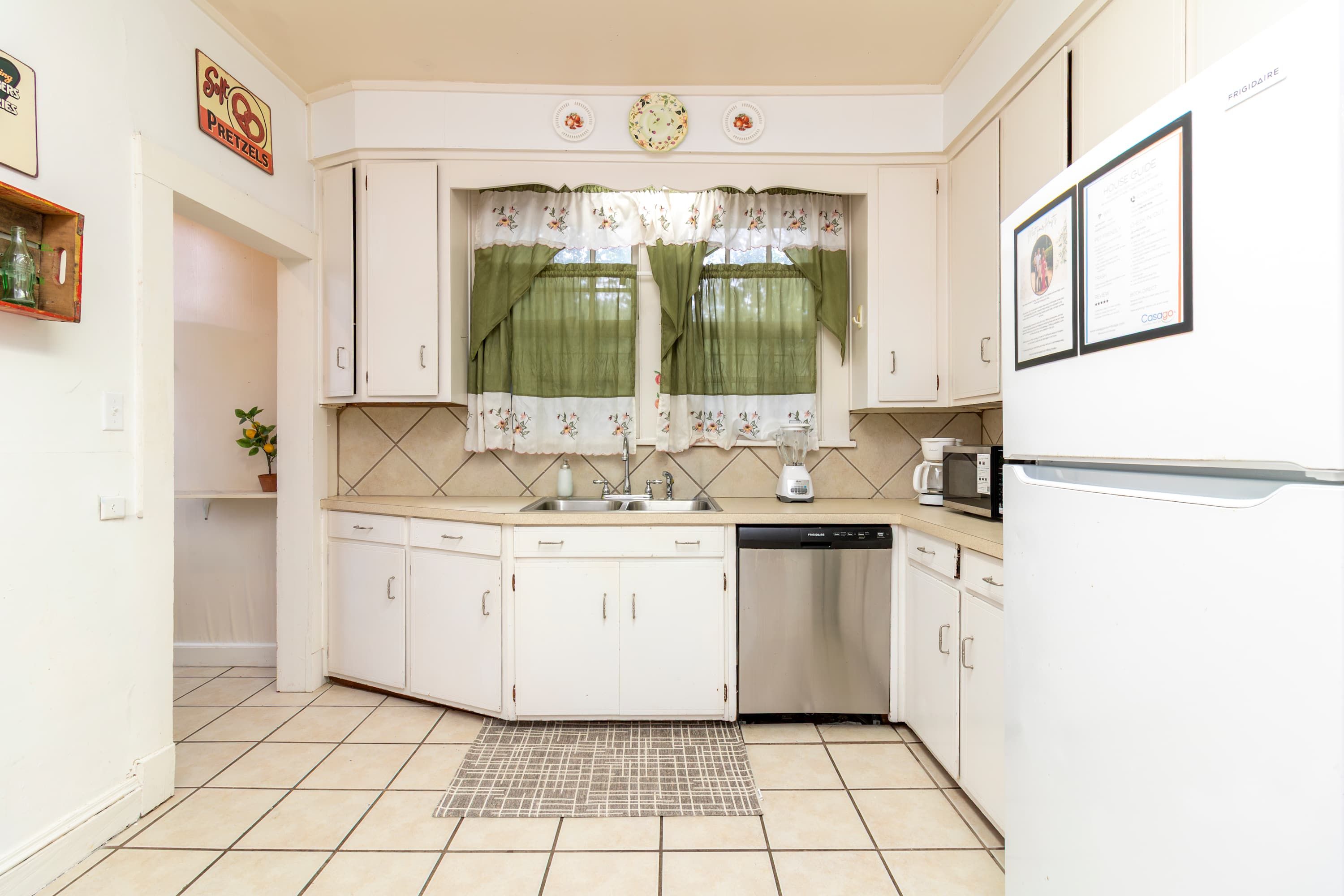 A compact kitchen with white cabinets, a built-in dishwasher, a microwave, and a window with floral curtains.