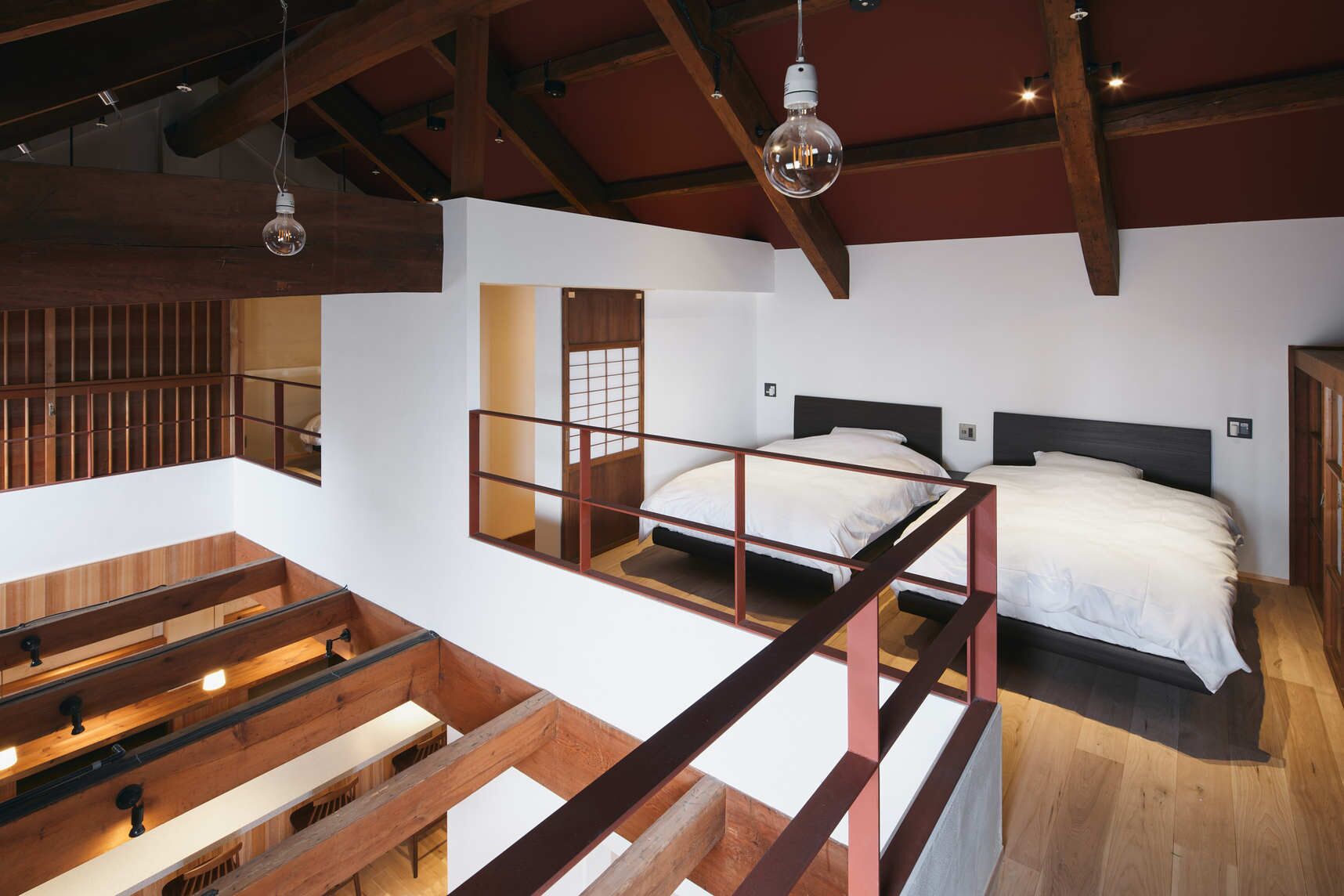 A loft-style bedroom with a high ceiling, exposed wooden beams, a mezzanine with a desk, and two beds with white bedding.