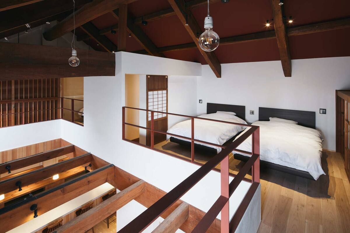 A loft-style bedroom with a high ceiling, exposed wooden beams, a mezzanine with a desk, and two beds with white bedding.