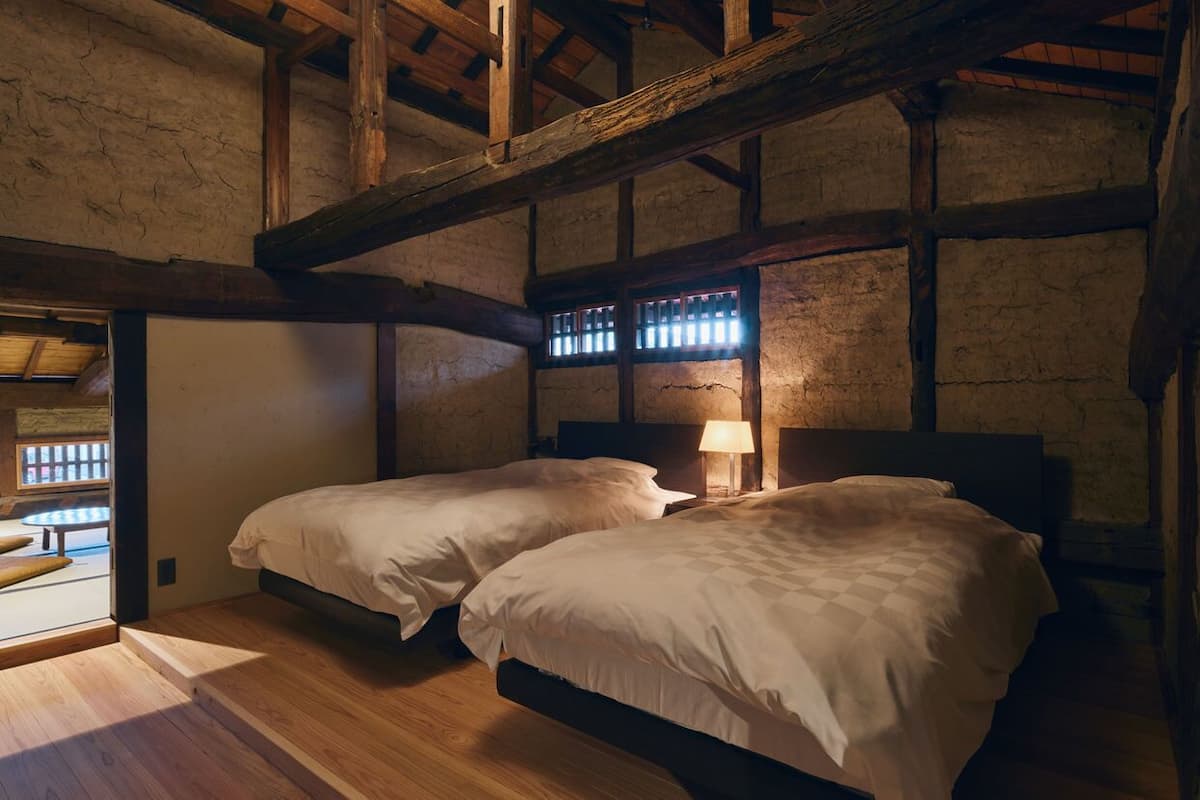 A rustic room with two beds, wooden beams, and a window with bars.