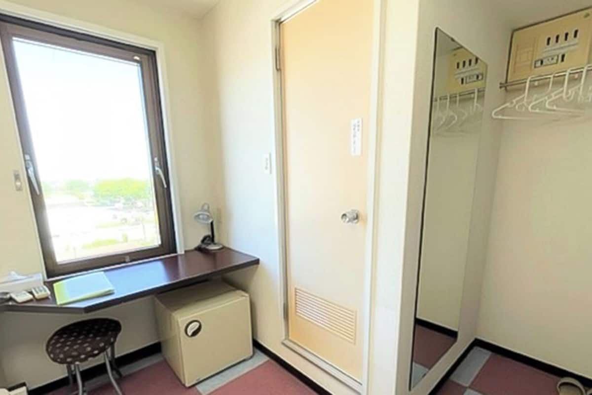 A small room with a desk, chair, window, and a wardrobe with hanging clothes.