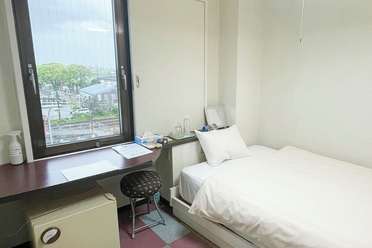 A hotel room with a bed, desk, chair, and a window with a view of buildings and trees.