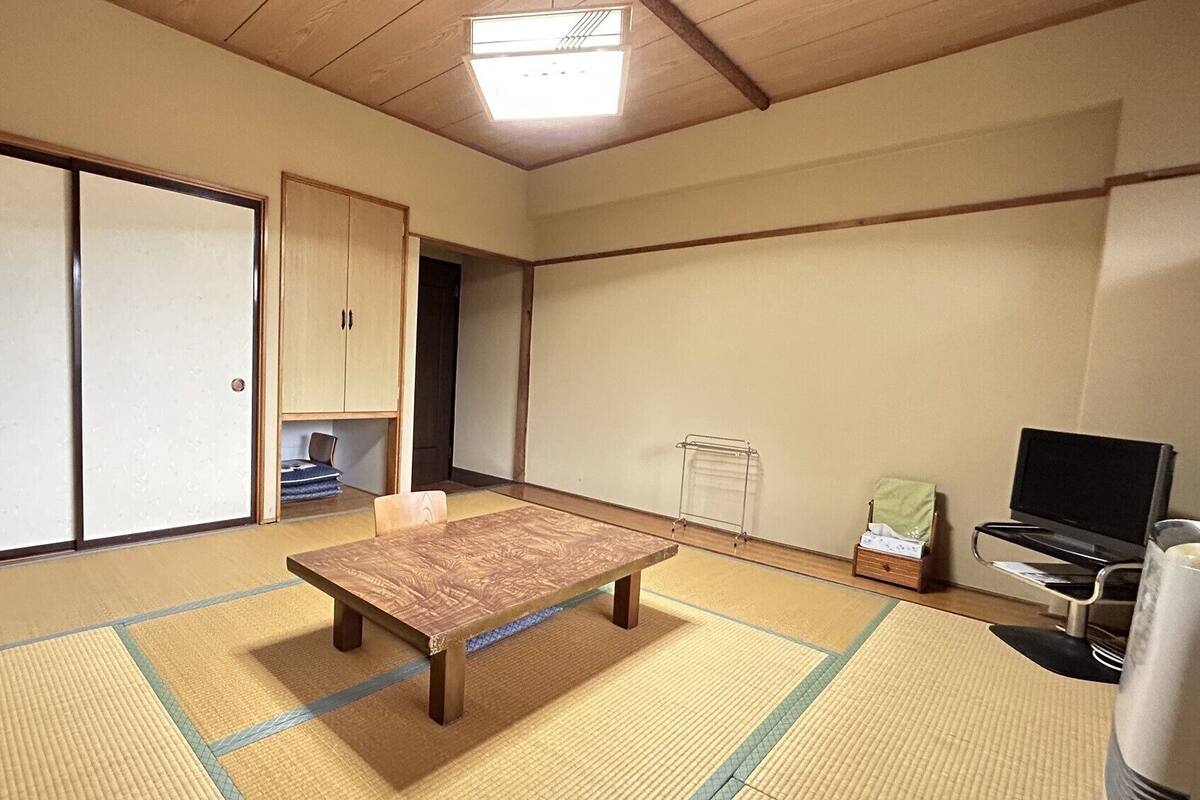 Una habitación japonesa tradicional con suelo de tatami, una mesa de madera baja, puertas correderas, un armario, un televisor y una pequeña estantería.
