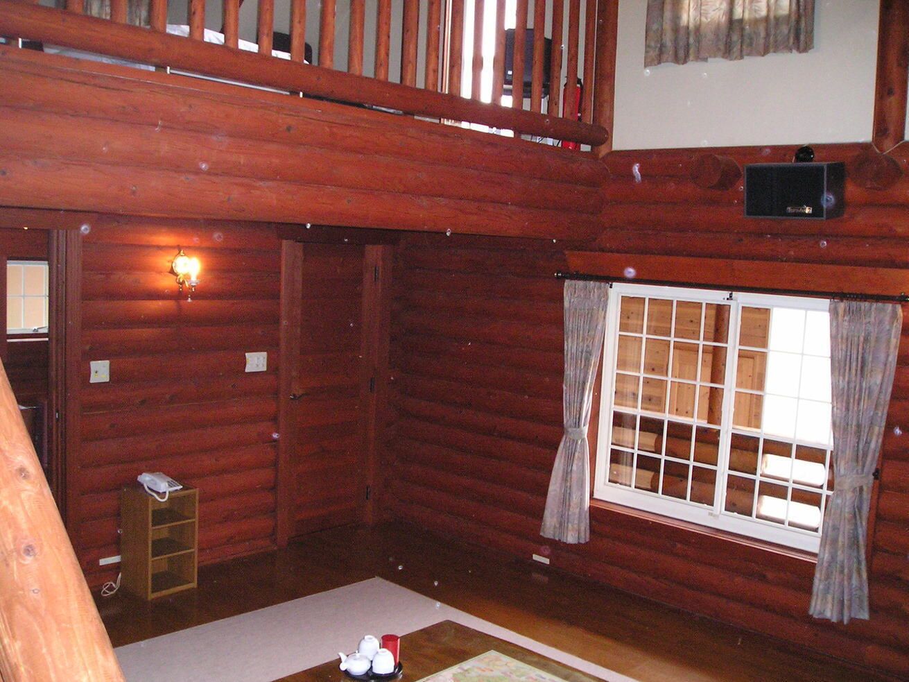 Een blokhutkamer met houten plafond, een raam met gordijnen, een klein houten plankje en een aan de muur bevestigde thermostaat.