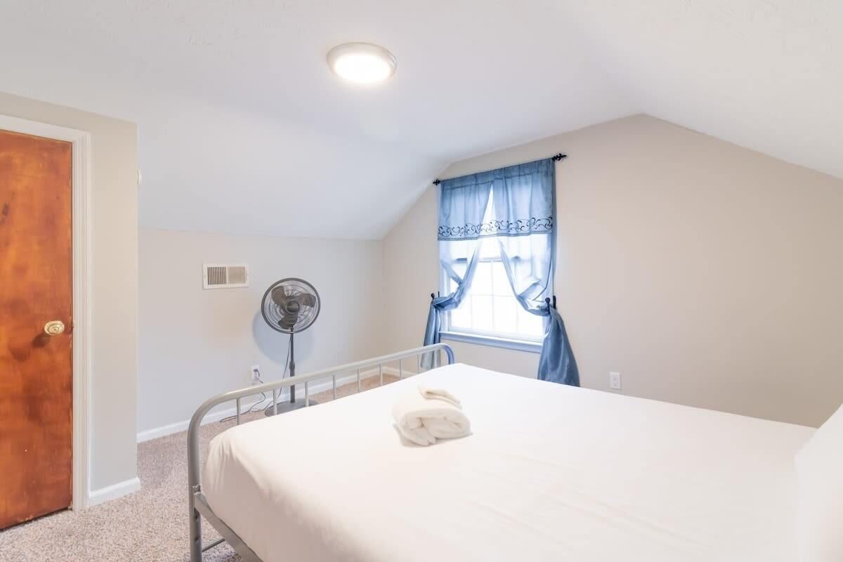 A bedroom with a bed, a bedside fan, a window with curtains, and a door.