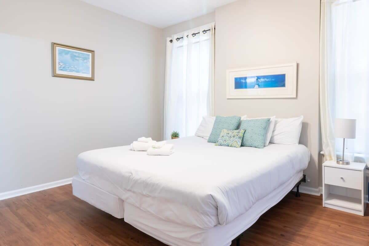 Une chambre à coucher avec un lit, une table de chevet, une fenêtre avec des rideaux et un tableau encadré au mur.
