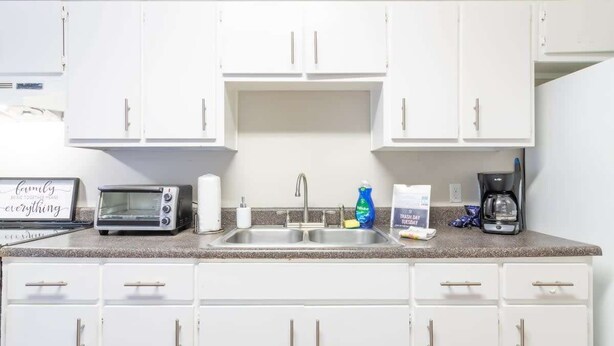 A kitchen with white cabinets, a double sink, a toaster, a coffee maker, and a trash can.