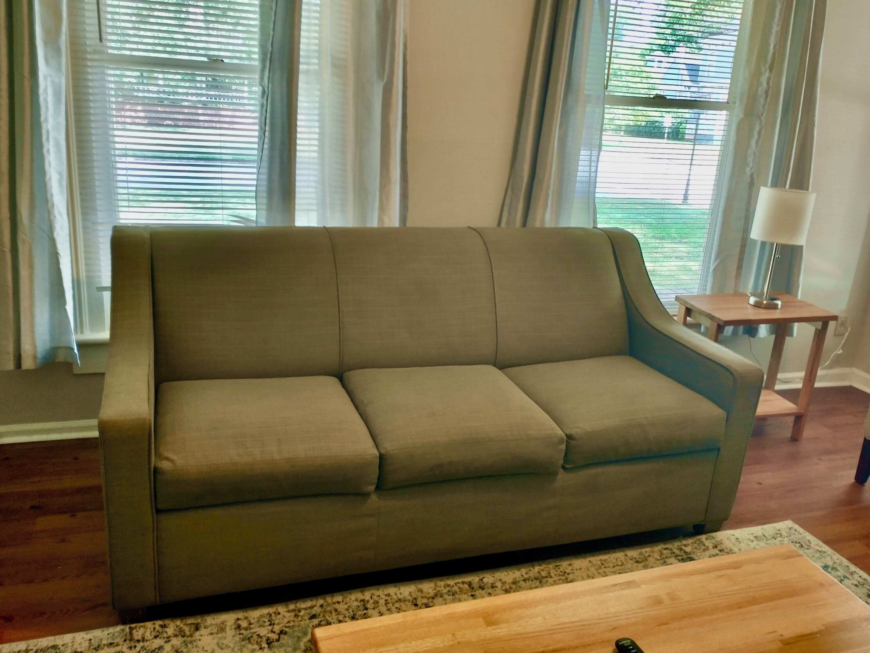 A beige sofa in a room with wooden flooring, a small wooden side table with a lamp, and a view through large windows with curtains.