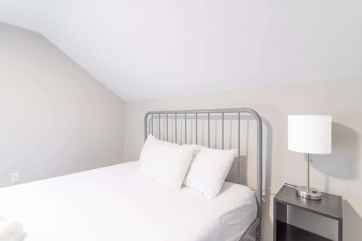 A single bed with a metal headboard, a bedside table with a lamp, and a plain white wall.