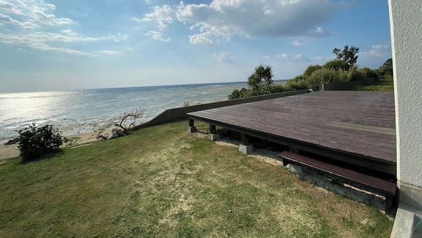A deck with a view of the ocean, a grassy area, and a white wall. - Golden Mile Villa (Amami)