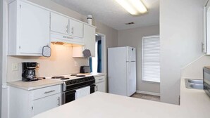 Une cuisine de taille réduite, avec des meubles blancs, une cuisinière, un réfrigérateur et une fenêtre équipée de stores.
